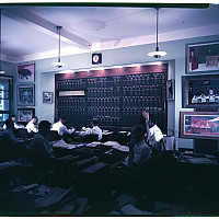 Archival color photograph of an office with gentlemen working at rows of desks facing a Wall Street-style trading board flanked by artworks.