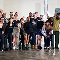 A group of dance students standing side-by-side in a museum gallery with pieces of an artwork visible in the background