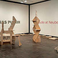 Gallery space with modular wooden sculptures arranged on a brown tile floor. Pieces of unassembled wood lie on the floor. The wall text read