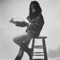 A person wearing a gorilla mask, sweater, fishnet stockings, and boots sits on a stool, crossing their legs and holding a peeled banana, poi