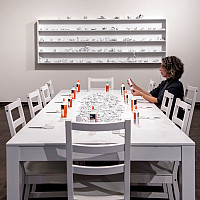 Woman seated at a long white table covered with broken pieces of white ceramic cups, white twine, bottles of white glue, and white scissors.