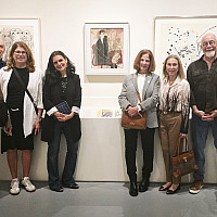 A group of thirteen casually dressed adults stands smiling in an art gallery, posing in front of three framed artworks displayed on a wall.