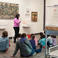 A group of students seated on the floor listing to a Volunteer Museum Educator describe one of Jackson Pollock's paintings