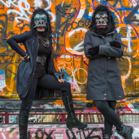 Two members of the artist collective The Guerrilla Girls in black clothing and gorilla masks pose in front of a wall covered in graffitti