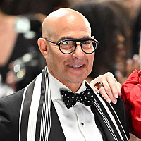 A bald man in a tuxedo with glasses stands next to a woman with short white hair in a red dress; both are smiling at a formal event.