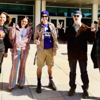 Five adults stand smiling in front of a modern building, wearing colorful, eclectic, and retro-inspired outfits including bright patterns, s