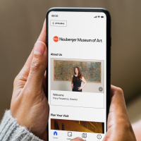 A person holds a smartphone displaying the Neuberger Museum of Art app, showing a welcome message from the director and navigation icons at