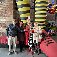 Four adults stand smiling in front of a whimsical, oversized inflatable sculpture with black and yellow striped legs. The group holds hands,