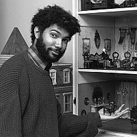 A young Fred Wilson, with curly hair and beard, wearing a sweater, gestures toward an elaborate multi-room dollhouse filled with furniture