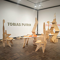 Gallery space with modular wooden sculptures arranged on a brown tile floor. Pieces of unassembled wood lie on the floor. The wall text read