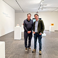 Two men stand smiling in a modern art gallery with white walls, surrounded by geometric sculptures displayed on white pedestals. The word TR