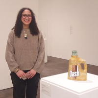 A person stands in a museum next to a sculpture resembling a yellow Dash detergent bottle. Several other sculptures are displayed on white