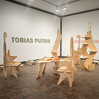Gallery space with modular wooden sculptures arranged on a brown tile floor. Pieces of unassembled wood lie on the floor. The wall text read