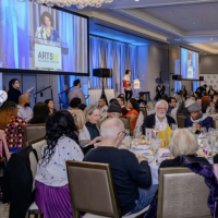 A large group of people sit at round tables in a banquet hall, listening to a speaker at a podium. The event is formal, with a projected scr