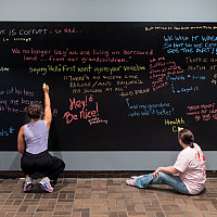 Three women in casual clothing writing with colored chalk on a large black chalkboard