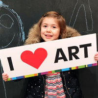 A smiling young child stands in front of a chalkboard with drawings, holding a sign that reads I ? ART with a red heart symbol. The child we