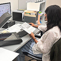 Student with dark long hair, white blouse, and jeans sits at an office desk with a book, desktop computer, and scanner.