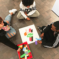 Three people sit on a tiled floor arranging colorful paper shapes around a sheet of abstract art, working together in a creative activity.