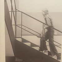 A black-and-white photo shows a person walking up a staircase indoors, with one hand on the railing. The image is slightly blurred.