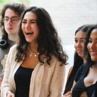 A group of young adults stand together, smiling and laughing. One woman in a white blouse is laughing loudly, while others look at each othe