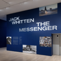 A museum gallery with deep blue walls displays JACK WHITTEN THE MESSENGER in large white letters surrounded by black-and-white photos; a lar