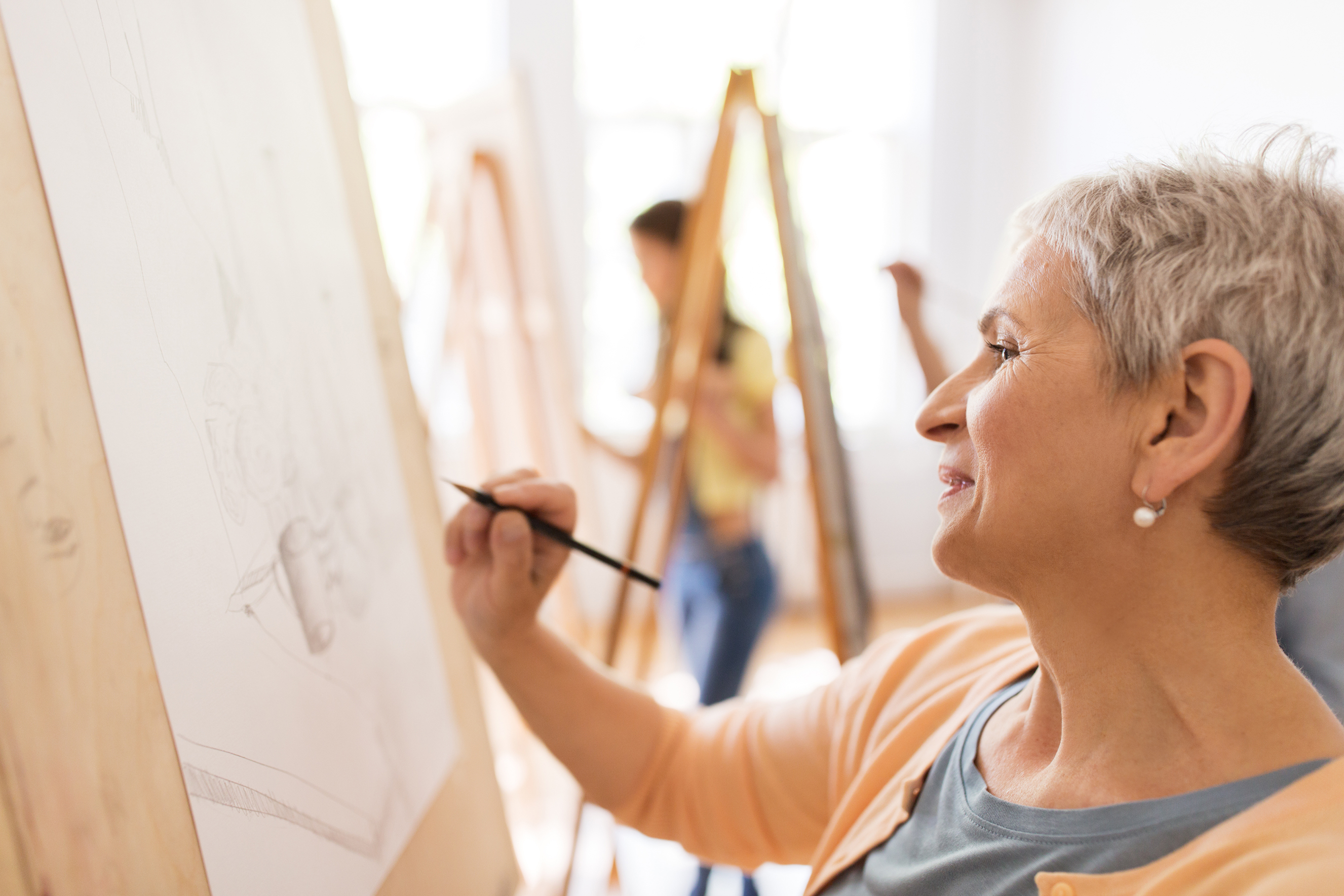 art school, creativity and people concept - happy senior woman artist with easel and pencil drawing picture at studio