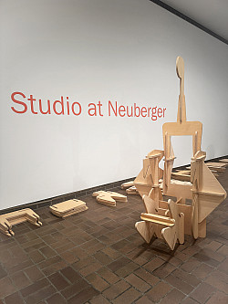A wooden abstract sculpture sits on a brown tiled floor in front of a white wall with the words Studio at Neuberger in large red letters. Ad