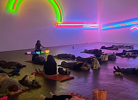People lie on mats in a dimly lit room, eyes closed, while a woman sits at the front near glowing neon lights on the wall, possibly leading