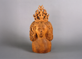 A sculpted bust of a woman with intricate curly hair and a large, ornate crown, set against a plain gray background.