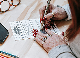 A person with long red nails and several rings draws abstract black lines on a white card, surrounded by colored pencils, glasses, a phone