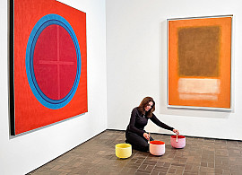 Woman seated on a brick floor with three crystal singing bowls. Behind her are two large paintings.