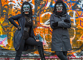 Two women in black outfits and gorilla masks pose confidently in front of a vibrant, graffiti-covered wall. One stands with a hand on their