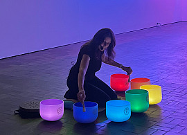 A woman plays colorful crystal singing bowls on the floor of a dark gallery, with bright neon light art in green, blue, yellow, and pink arc