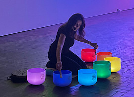 A woman plays colorful crystal singing bowls on the floor of a dark gallery, with bright neon light art in green, blue, yellow, and pink arc