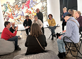 A group of people sits in a circle on chairs and bean bags in an art gallery, engaged in discussion. Colorful paintings are displayed on the