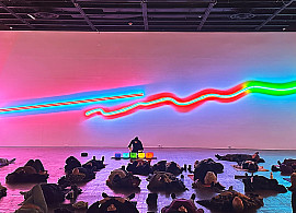 People lie on mats in a dimly lit room, eyes closed, while a woman sits at the front near glowing neon lights on the wall, possibly leading