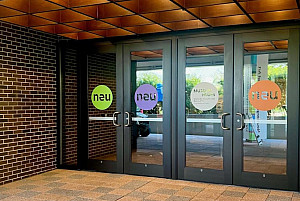 Photo of the entrance to the Museum featuring four glass doors, each with a decal of the letters neu centered in a color circle.