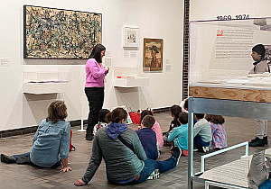 A group of students seated on the floor listing to a Volunteer Museum Educator describe one of Jackson Pollock's paintings