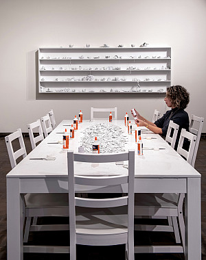 Woman seated at a long white table covered with broken pieces of white ceramic cups, white twine, bottles of white glue, and white scissors.
