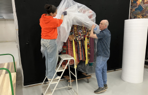 A young female on a ladder with a man standing, each holding part of a sheet of protective plastic that is being draped over a sculpture.