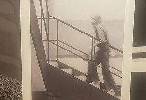 A black-and-white photo shows a person walking up a staircase indoors, with one hand on the railing. The image is slightly blurred.