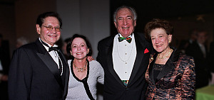 Four adults dressed in formal evening attire stand together and smile at the camera at a social or gala event, with a blurred background.