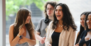 A group of young adults stand together, smiling and laughing. One woman in a white blouse is laughing loudly, while others look at each othe