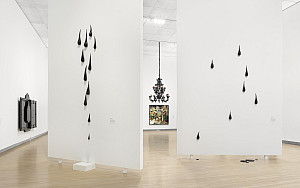 Contemporary art gallery with white walls, featuring black teardrop-shaped sculptures mounted at varying heights and a black chandelier in t