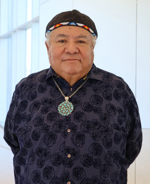 An older man stands indoors wearing a patterned dark shirt, a beaded cap, and a large turquoise necklace, with his hands behind his back and