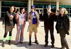 Five adults stand smiling in front of a modern building, wearing colorful, eclectic, and retro-inspired outfits including bright patterns, s