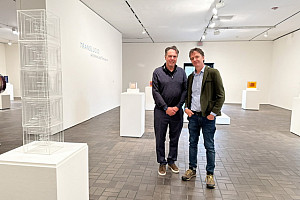 Two men stand smiling in a modern art gallery with white walls, surrounded by geometric sculptures displayed on white pedestals. The word TR