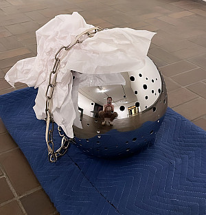 A woman is reflected in a shiny metal sphere with small holes sits on a blue padded mat. A crumpled paper protects the surface.