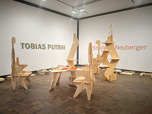 Gallery space with modular wooden sculptures arranged on a brown tile floor. Pieces of unassembled wood lie on the floor. The wall text read