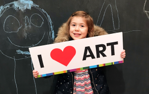 A smiling young child stands in front of a chalkboard with drawings, holding a sign that reads I ? ART with a red heart symbol. The child we